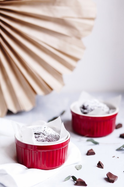 Chocolademuffins in rode kopjes. Kleine geglazuurde keramische ramekin met bruine cakes op een grijze en witte achtergrond.