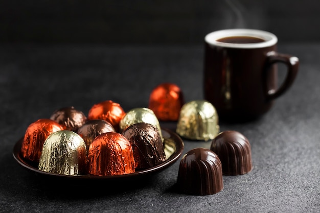 Chocoladesuikergoed verpakt in veelkleurige folie op een bord en kop warme koffie op zwarte achtergrond met kopieerruimte