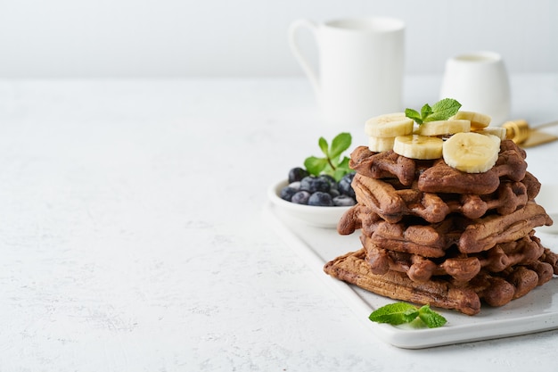 Chocolate banana waffles with maple syrup on white table, copy space, side view.
