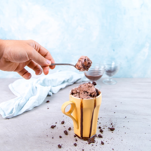 Chocolate brown ice cream in a regular yellow mug and spoon, on blue, with topping and crumbs. 