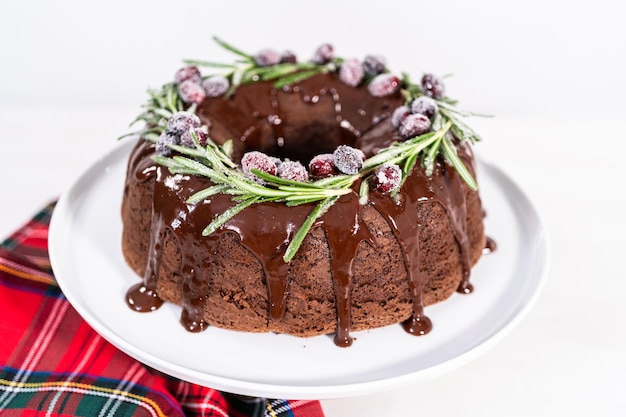 Chocolate bundt cake with chocolate frosting decorated with fresh cranberries and rosemary covered in a white sugar.