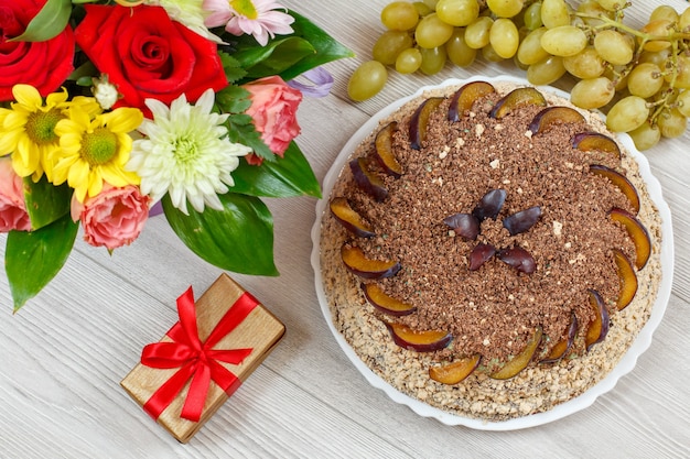 Chocolate cake decorated with plums, a bouquet of flowers, grapes and a gift box on the gray wooden boards.