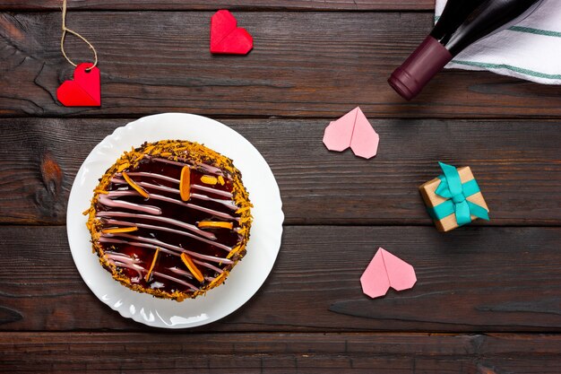 Chocolate cake, small paper hearts and a bottle of wine on a dark wooden table.