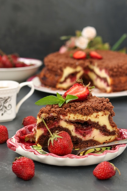 Chocolate cake with cottage cheese with strawberries is located on a dark surface, a piece of cake is located in the foreground on a plate