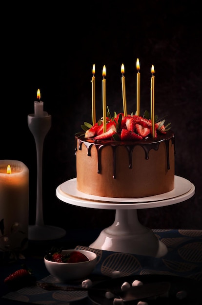 A chocolate cake with strawberries on it and lit candles on a white cake stand.