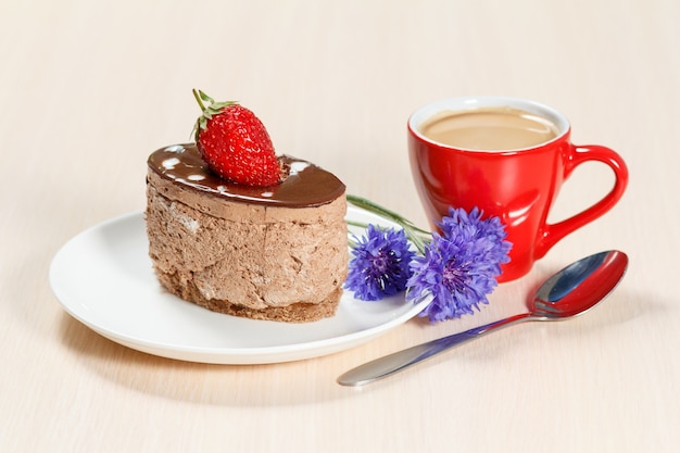 Шоколадный торт с клубникой на белой тарелке, васильками и чашкой кофе на деревянном столе