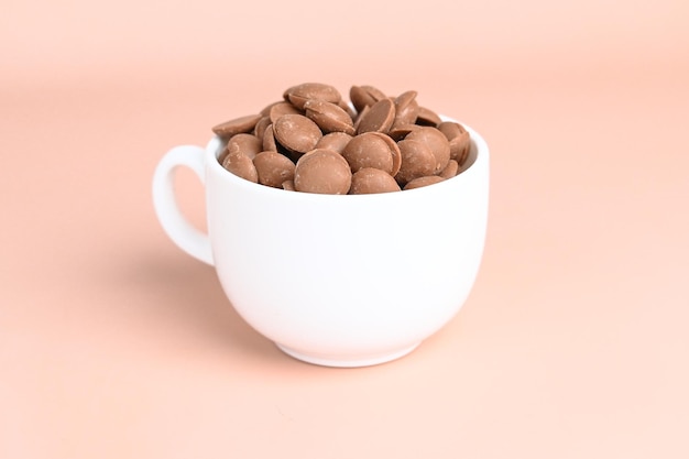 Chocolate callets in a white cup on a pastel background Tempered chocolate confectionery concept