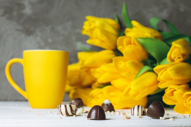 Chocolate candies, cup and bouquet of yellow tulips on white wooden surface on gray surface