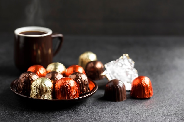 Фото Шоколадные конфеты, завернутые в разноцветную фольгу на тарелке и две чашки горячего кофе на черном с копией пространства