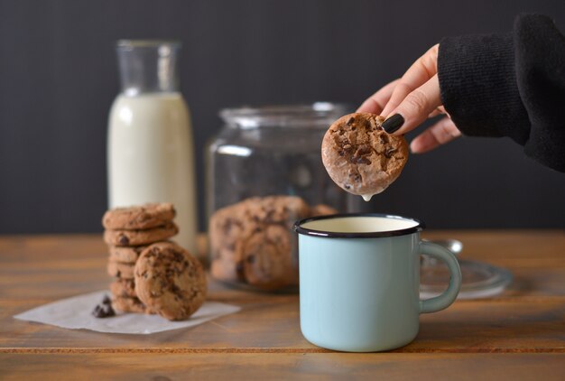chocolate chip cookies in glass jar with glass bottle of milk and turquoise enamel mug on wooden rustic background with women hand holding one cookie 