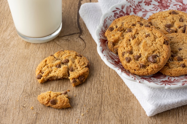 Chocolate chip and vanilla cookies on a decorated plate over textile with broken cookie and a glass of milk over a wooden table.