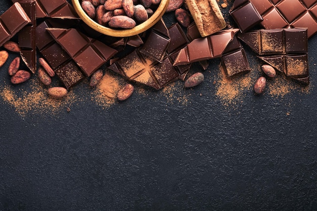 Chocolate composition of cocoa powder grated and bean cocoa\
bars and pieces of different milk and dark chocolate on black\
background baking chocolate texture top view with copy space mock\
up