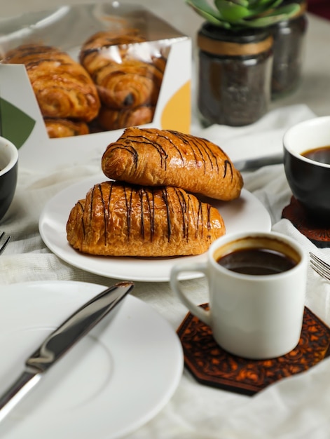 Chocolate Croissant served on plate with cup of black coffee with knife and fork isolated on napkin side view of french breakfast baked food item on grey background