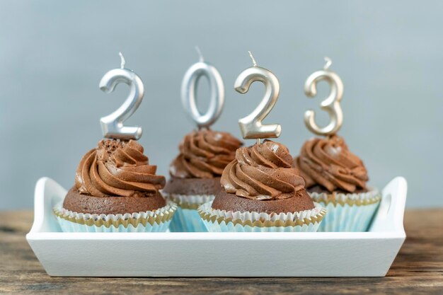 Chocolate cupcakes with buttercream frosting with silver 2023 candles on wooden table on light concrete background