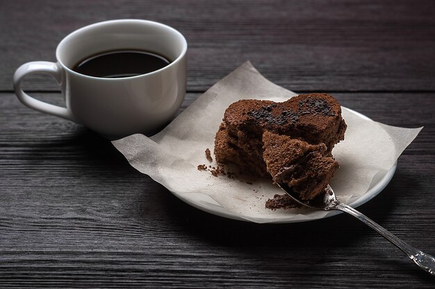 Chocolate dessert in the form of a heart on a white saucer next to a cup of fragrant coffee stands on a wooden old background