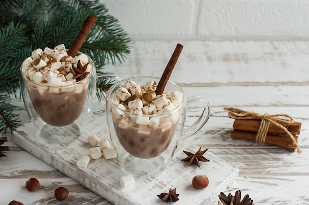 Bevanda al cioccolato in tazza trasparente con marshmallow, bastoncini di cannella e stelle di anice su uno sfondo di legno bianco di fronte al muro di mattoni.