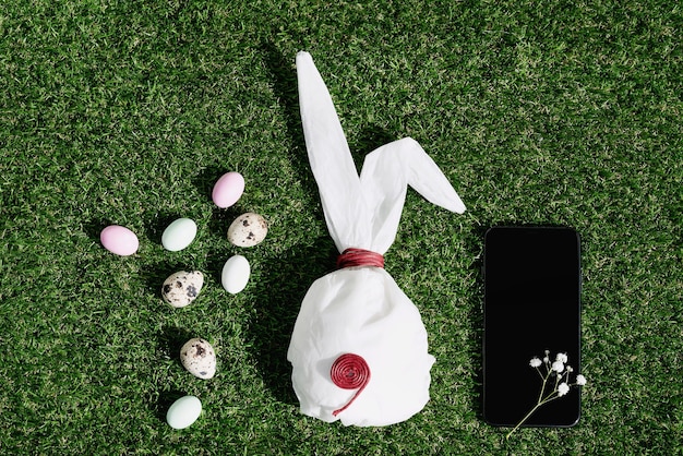 Chocolate Easter eggs glazed with pastel coloring and quail eggs over green grass. Bunny shaped white baking paper. Black cell phone. Happy Easter concept. Copy space. Top view