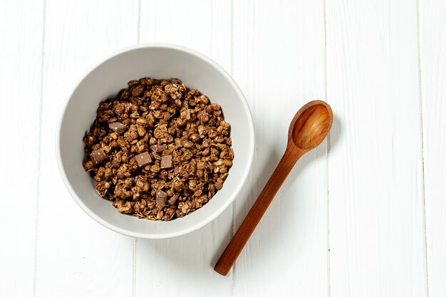 Chocolate granola in a white bowl in a composition with a spoon on white wooden background. 