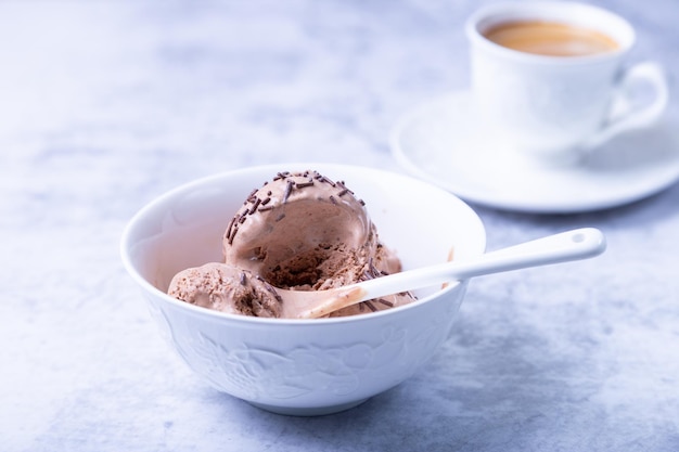 Chocolate ice cream with chocolate sprinkle in a white bowl on a light background. Three balls of homemade ice cream and a cup of coffee. Close-up.