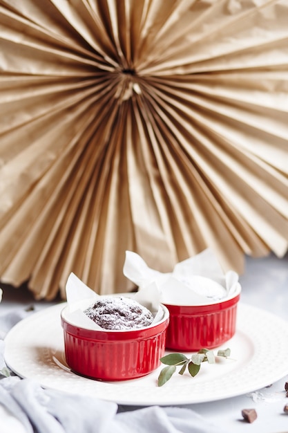 Фото Шоколадные кексы в красных чашках. маленькая глазированная керамическая формочка с коричневыми лепешками на серо-белом фоне.