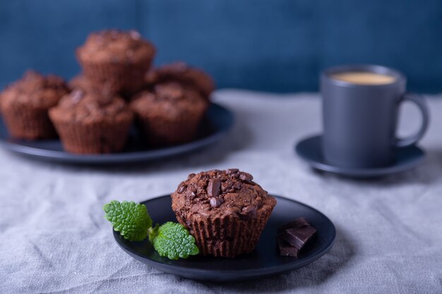 黒い皿にミントとチョコレートのマフィン。自家製のベーキング。背景には一杯のコーヒーとマフィンのプレートがあります。大理石のテーブルと青い背景。