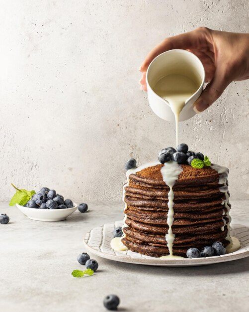 Chocolate pancakes with white sauce and berries, the hostess pours the milk jug sauce. Light textured background, breakfast concept, cooking. Vertical orientation
