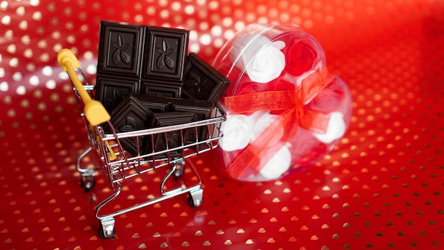 Foto cioccolato nel carrello della spesa e regalo di rose su sfondo rosso. 14 febbraio vendita. concept creativo minimal