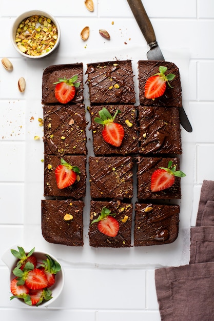 Chocolate squares with pistachio nuts and strawberries on white paper on a light surface, top view, horizontal composition. Flat food.