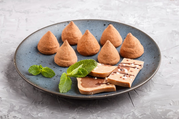 Tartufo al cioccolato con un pezzo di cioccolato al latte sul piatto in ceramica blu su sfondo grigio cemento.