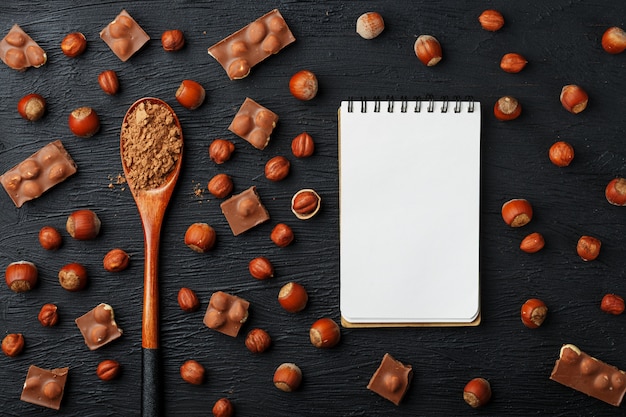 Photo chocolate with hazelnuts, a notebook with blank pages and a wooden spoon with cocoa, surrounded by nuts in the shell and peeled.