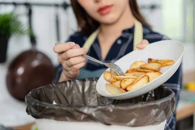 정크 식사의 콜레스테롤은 뚱뚱한 아시아 젊은 가정 여성이 부엌에 있는 감자 칩 스낵에서 음식을 쓰레기통에 긁어 내고 환경적으로 책임 있는 생태학입니다.
