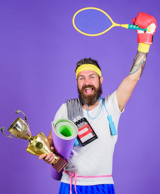 Choose favorite sport. Sport concept. On way to achievement. Sport shop assortment. Man bearded athlete hold sport equipment jump rope fitness mat boxing glove expander racket and golden goblet.