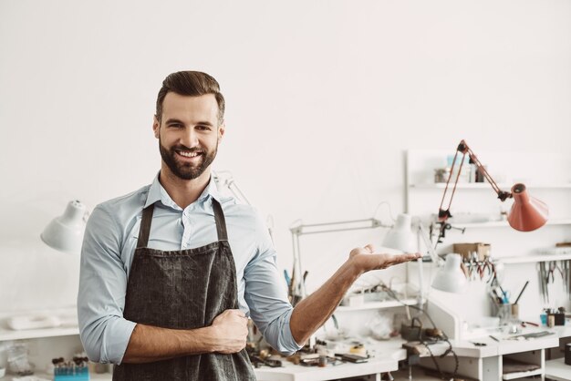 Choose it. Portrait of cheerful male jeweler wearing apron gesturing while standing in his jewelry making studio. Business. Jewelry workshop