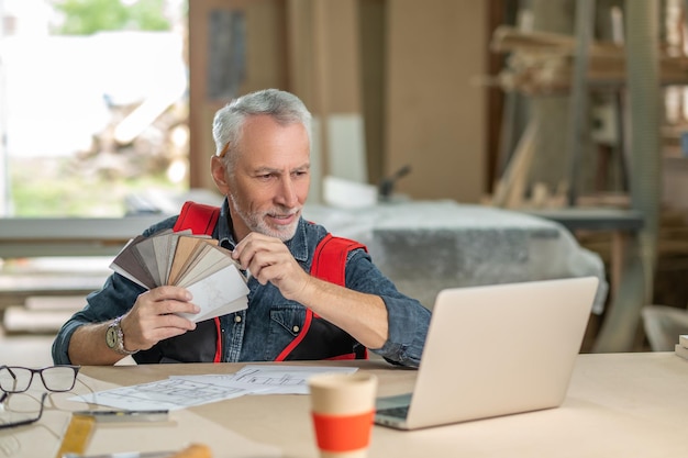 Choosing colors. Bearded gray-haired man sitting at the table with a colors palette in hands