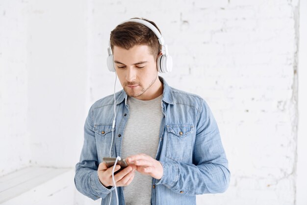 Choosing track. Confident brunette wearing headphones and bowing head while using his gadget