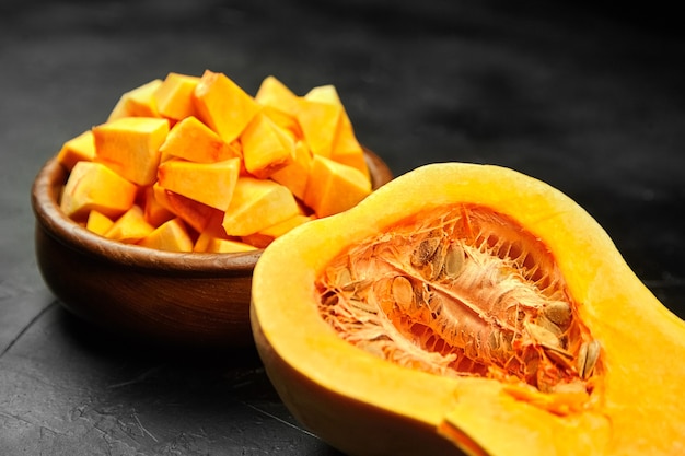 Chopped butternut squash, pumpkin half, fresh vegetable pieces in wooden bowl on black stone table, selective focus. Food ingredient