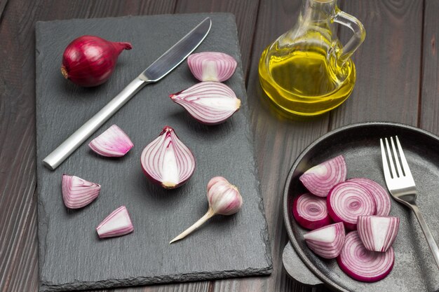 Photo chopped onion, knife and one whole onion on cutting board. chopped onions and fork in frying pan. oil bottle. dark wooden background. top view