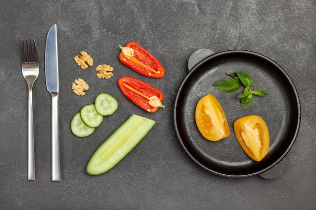 Chopped yellow tomatoes in frying pan. Cucumber and slices of red pepper, fork and knife on table. Black background. Flat lay.