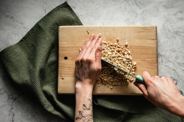 Chopping peanut with a knife on a wooden cutting board, a green linen towel, marble backdrop, top view horizontal photo. Cookie recipe.