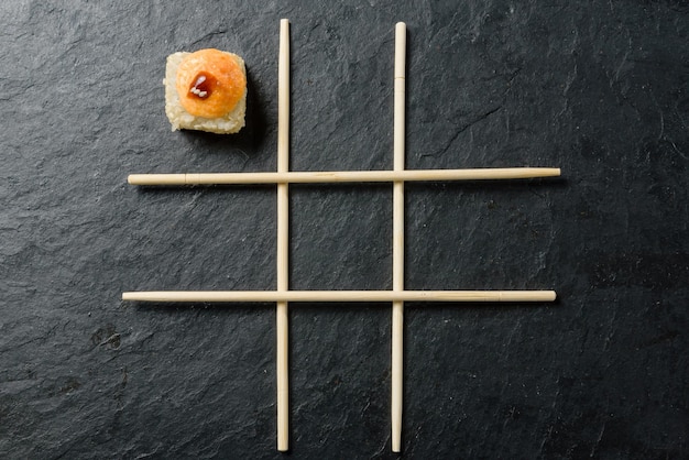 Chopsticks arranged in a tictactoe pattern and one roll on a dark stone background