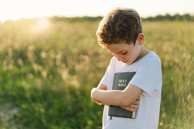 Il ragazzo cristiano tiene la bibbia nelle sue mani leggendo la sacra bibbia in un campo durante il bel tramonto concetto di fede, spiritualità e religione speranza di pace