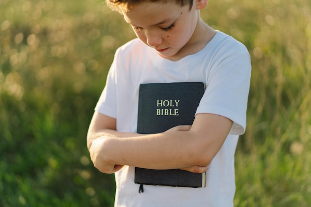 Christian boy holds bible in her hands reading the holy bible\
in a field during beautiful sunset concept for faith spirituality\
and religion peace hope