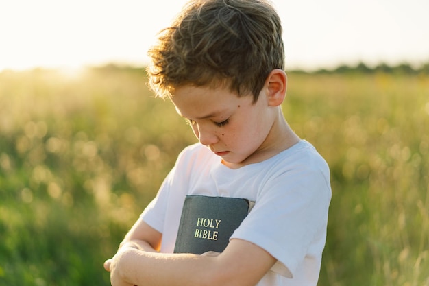 Christian boy holds bible in her hands reading the holy bible\
in a field during beautiful sunset concept for faith spirituality\
and religion peace hope