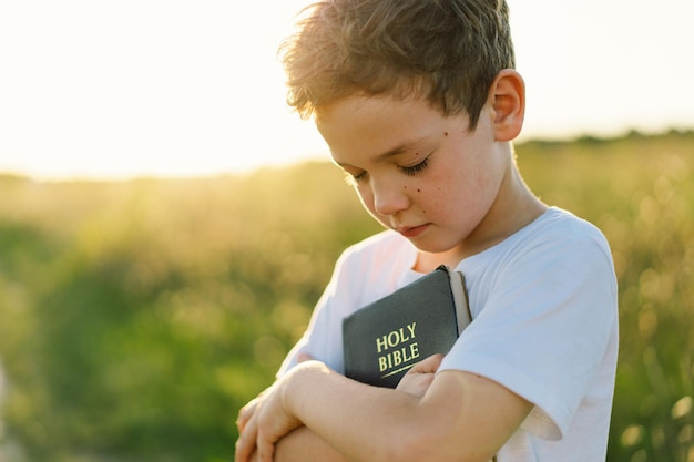 Il ragazzo cristiano tiene la bibbia nelle sue mani leggendo la sacra bibbia in un campo durante il bel tramonto concetto di fede, spiritualità e religione speranza di pace