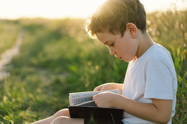 Christian boy holds bible in her hands reading the holy bible\
in a field during beautiful sunset concept for faith spirituality\
and religion peace hope