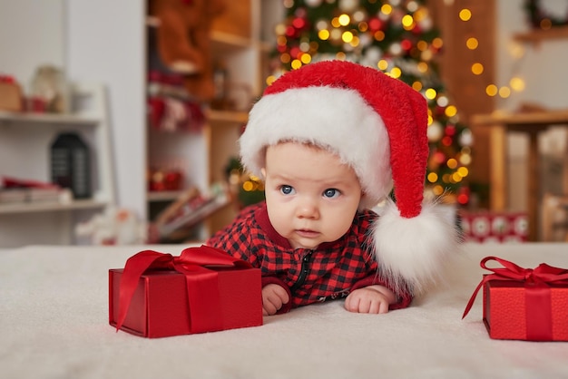 Christmas baby boy in santa hat on background of christmas tree with gifts.