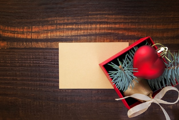 Christmas background: red gift box with a Christmas toy in the shape of a heart, a golden bell , fir branches and card for congratulatory text