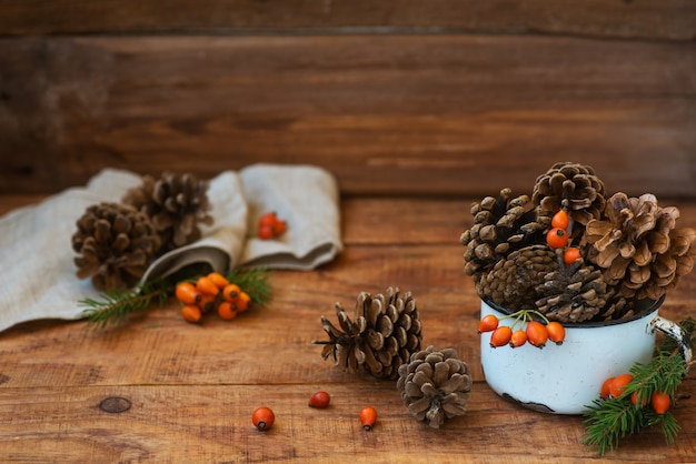 Christmas background in rustic style. Atmospheric composition of pine cones in vintage cup, rosehips berries, spruce branches and wheat spikelets is placed on wooden surface. CopySpace, Flat lay