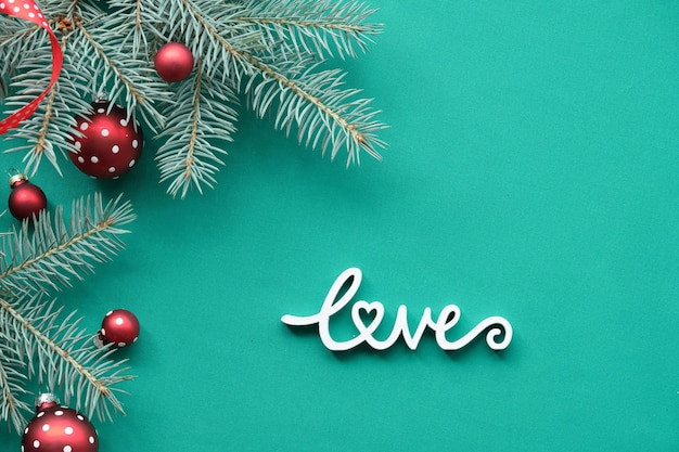 Christmas background on turquoise, green textile with wooden text Love. Top view on fir twigs decorated with red glass baubles, traditional Xmas toys.