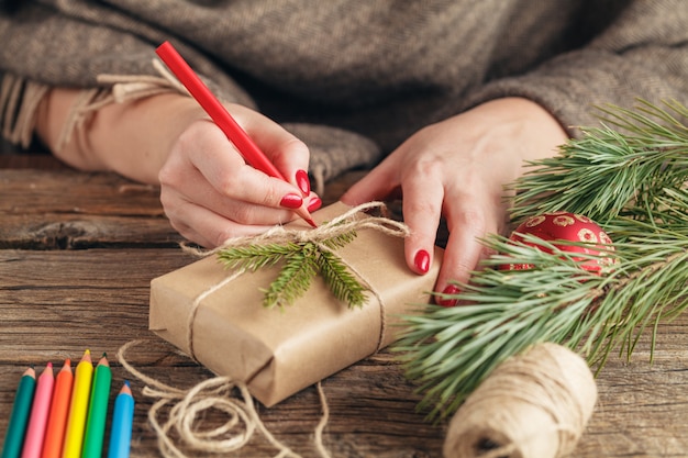 Christmas background. view of hands writing on gift box. Packed gifts and scrolls, spruce branches on shabby wooden table. Workplace for preparing handmade new year decorations.